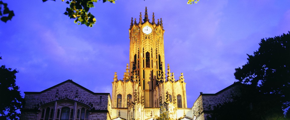 Illuminated Gothic clock tower against a purple twilight sky, framed by tree silhouettes